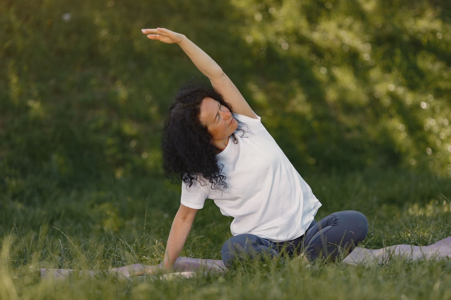 Mature woman doing seated side bend yoga pose in a sunny park.