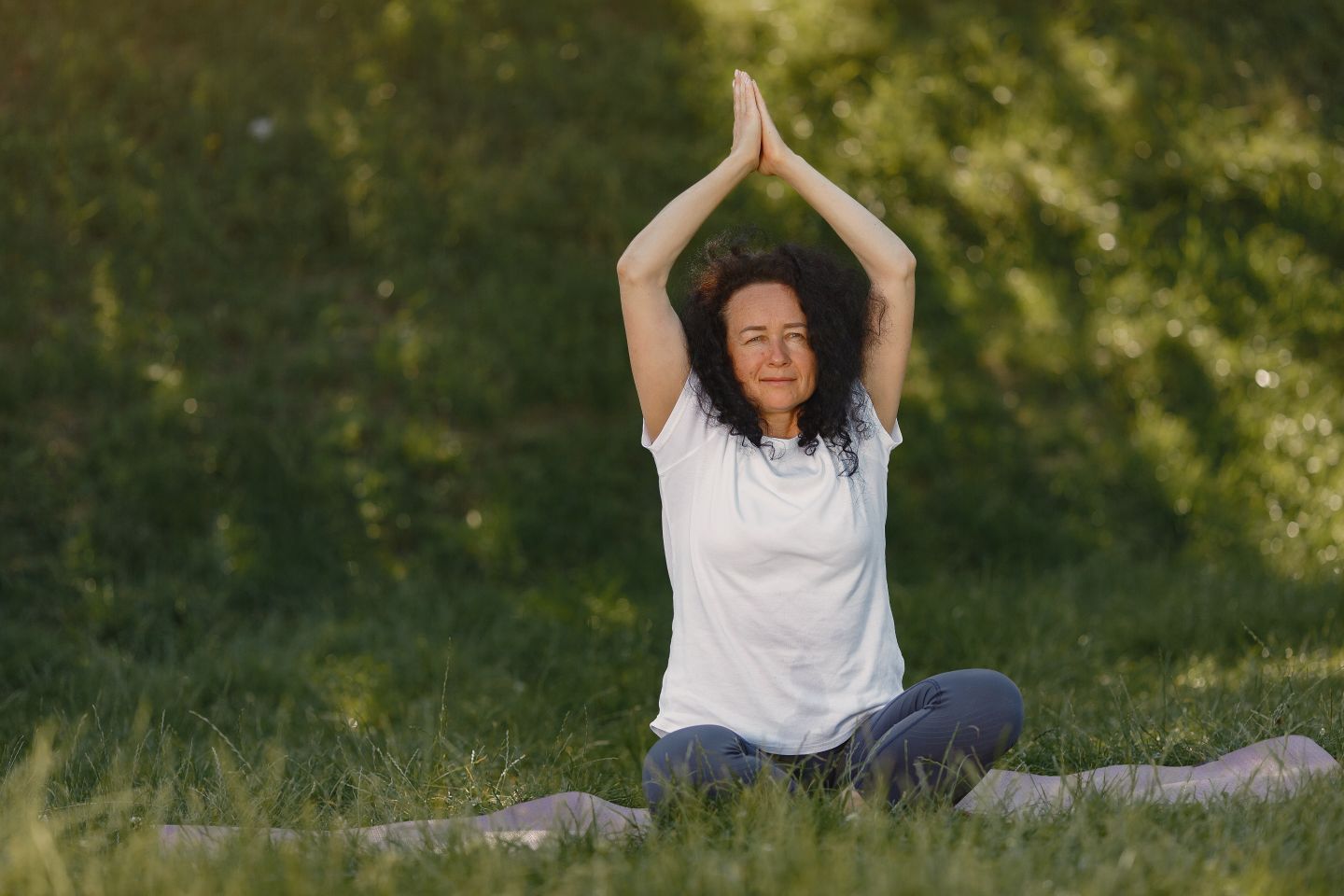 Adult woman doing yoga in a seated pose with hands raised on a grassy outdoor setting.